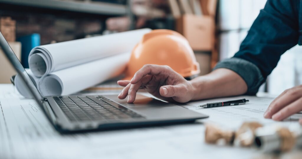 uomo al computer con caschetto e progetti di un edificio